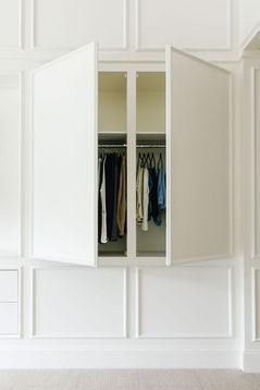 A sleek and minimal built-in wardrobe integrated into paneled white walls, featuring open doors revealing neatly hung clothing. The design blends seamlessly with the surrounding decor, showcasing functionality and elegance. Designed by Bar Kay Design Co., an interior design business located in Oregon, specializing in services across Eugene, Bend, and Portland, showcasing high-quality craftsmanship and innovative design.