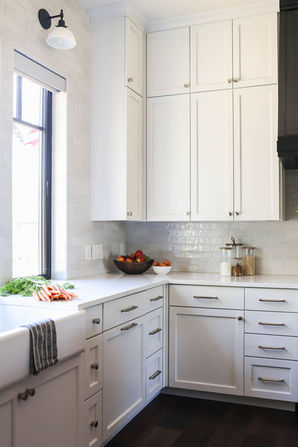 A bright and clean kitchen corner featuring white cabinetry with sleek hardware and a light subway tile backsplash. A farmhouse sink with a striped towel is positioned under a window, bringing in natural light. The countertop is styled with a bowl of fresh produce, glass jars, and freshly washed carrots for a natural and inviting touch. The dark wood flooring contrasts beautifully with the light tones, creating a warm and sophisticated aesthetic. Designed by Bar Kay Design Co., an interior design business located in Oregon, specializing in services across Eugene, Bend, and Portland, showcasing high-quality craftsmanship and innovative design.