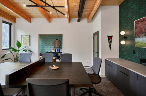 A contemporary office space featuring a large dark wood conference table, ergonomic chairs, and exposed wooden ceilings with modern lighting. The design incorporates a mix of textures, including a green accent wall, dark cabinetry, and subtle decorative details, blending functionality with aesthetic appeal. Designed by Bar Kay Design Co., an interior design business located in Oregon, specializing in services across Eugene, Bend, and Portland, showcasing high-quality craftsmanship and innovative design.