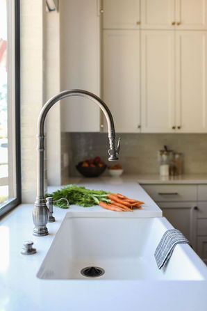 A close-up view of a farmhouse-style kitchen sink with a sleek gooseneck faucet set in a white countertop. Freshly washed carrots with green tops rest beside the sink, adding a natural and inviting touch. The background features white cabinetry and a light-colored backsplash, creating a clean and airy aesthetic. Designed by Bar Kay Design Co., an interior design business located in Oregon, specializing in services across Eugene, Bend, and Portland, showcasing high-quality craftsmanship and innovative design.