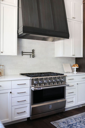 A modern kitchen featuring a stainless steel range with a sleek black range hood set against a light subway tile backsplash. The surrounding white cabinetry provides ample storage, while a pot filler faucet above the stove adds convenience. A small loaf of bread on a white pedestal stand and a decorative cookbook add a touch of warmth and personality to the clean and functional design. Designed by Bar Kay Design Co., an interior design business located in Oregon, specializing in services across Eugene, Bend, and Portland, showcasing high-quality craftsmanship and innovative design.