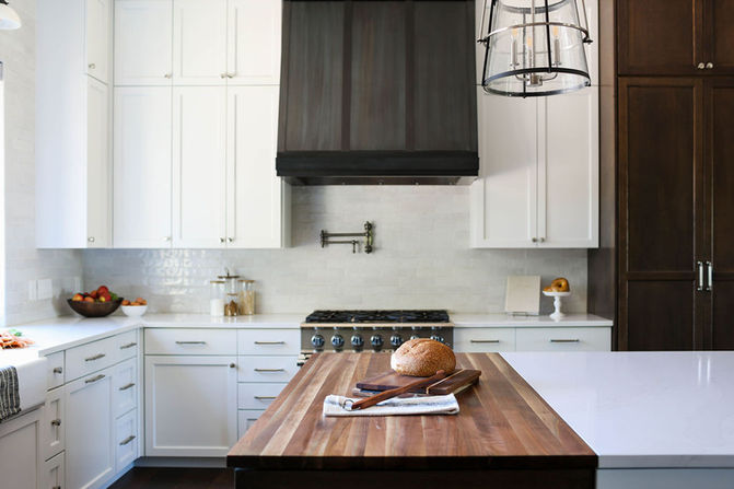 A beautifully designed kitchen with a large butcher block island as the centerpiece, featuring a loaf of bread, wooden utensils, and a folded cloth for a warm, inviting touch. The backdrop includes white cabinetry, a stainless steel range, and a sleek black range hood set against a light subway tile backsplash. A pot filler faucet above the stove adds functionality, while a modern pendant light with a metal frame hangs above the island. Designed by Bar Kay Design Co., an interior design business located in Oregon, specializing in services across Eugene, Bend, and Portland, showcasing high-quality craftsmanship and innovative design.