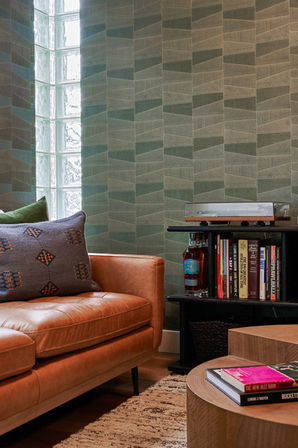 A cozy corner featuring a tufted tan leather sofa with patterned pillows, a modern bookshelf stocked with books and decor, and a textured green accent wall with glass block detailing. The space is complemented by a round wooden coffee table and a neutral area rug, creating a warm and inviting atmosphere. Designed by Bar Kay Design Co., an interior design business located in Oregon, specializing in services across Eugene, Bend, and Portland, showcasing high-quality craftsmanship and innovative design.