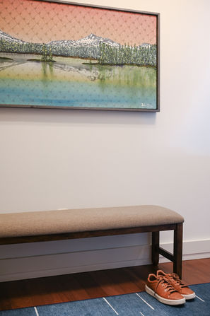 A cozy bench placed beneath a vibrant landscape artwork, paired with neatly positioned sneakers on a textured rug, creating a welcoming entryway. Designed by Bar Kay Design Co., an interior design business located in Oregon, specializing in services across Eugene, Bend, and Portland, showcasing high-quality craftsmanship and innovative design.