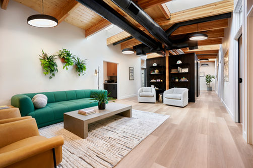 A bright and airy open-concept office space featuring a green sofa, a modern concrete coffee table, and a textured area rug. The design incorporates natural wood elements, black exposed ductwork, and floating planters for a fresh and contemporary feel. Cozy seating nooks with bookshelves add functionality and warmth. Designed by Bar Kay Design Co., an interior design business located in Oregon, specializing in services across Eugene, Bend, and Portland, showcasing high-quality craftsmanship and innovative design.