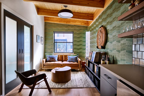 A modern lounge area featuring a tan leather sofa with patterned pillows, a round wooden coffee table, and a sleek black bookshelf displaying books and decor. The space is accented by a textured green wall, glass block windows, exposed wooden beams, and a neutral area rug, creating a stylish and inviting atmosphere. Designed by Bar Kay Design Co., an interior design business located in Oregon, specializing in services across Eugene, Bend, and Portland, showcasing high-quality craftsmanship and innovative design.
