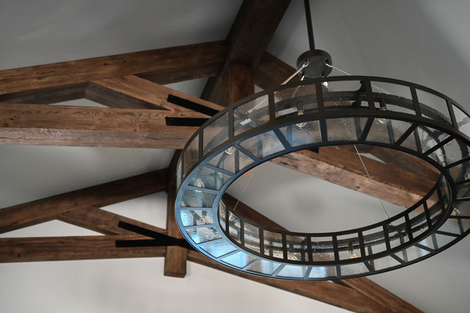 A circular metal chandelier with glass panels hangs from a wooden beam ceiling. Designed by Bar Kay Design Co., an interior design business located in Oregon, specializing in services across Eugene, Bend, and Portland, showcasing high-quality craftsmanship and innovative design.