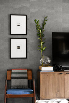 A stylish living area featuring framed abstract artwork on a textured wall, creating a sophisticated backdrop. A wooden chair with a blue cushion and a striped throw is positioned next to a modern media console. A sleek black vase with a simple plant adds a touch of greenery, while stacked books and a sculptural object on the console complete the balanced and elegant design. Designed by Bar Kay Design Co., an interior design business located in Oregon, specializing in services across Eugene, Bend, and Portland, showcasing high-quality craftsmanship and innovative design.