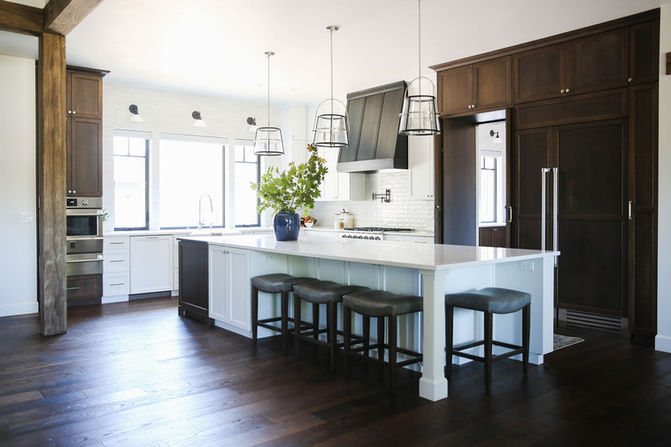 A beautifully designed kitchen featuring a large white island with upholstered bar stools and a blue vase of greenery as a centerpiece. The kitchen combines white cabinetry, a black range hood, and dark wood cabinets for a balanced and sophisticated look. Pendant lights with metal frames add modern elegance, while large windows allow natural light to brighten the space. The dark wood flooring ties the design together, creating warmth and contrast. Designed by Bar Kay Design Co., an interior design business located in Oregon, specializing in services across Eugene, Bend, and Portland, showcasing high-quality craftsmanship and innovative design.