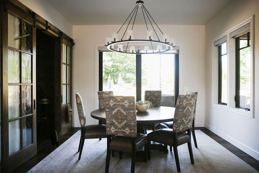 A bright and inviting dining room featuring a round wooden table surrounded by upholstered chairs with a tribal-inspired pattern. A large circular chandelier with a modern design hangs above the table, providing elegant lighting. The space includes large windows that bring in natural light and a sliding barn door on one side, adding a rustic yet contemporary touch. Designed by Bar Kay Design Co., an interior design business located in Oregon, specializing in services across Eugene, Bend, and Portland, showcasing high-quality craftsmanship and innovative design.