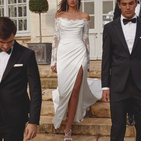 Charmeuse fit and flare gown with off-the-shoulder neckline and corded Alencon lace long sleeves. Sleeves finished with buttons at wrists. Features cowl detail at neckline with peek-a-boo lace. Flattering ruching throughout bodice continues onto top of skirt revealing a sexy slit. Corded Alencon lace continue straight across back neckline with charmeuse v-back with pleated detailing. Buttons down the back ending at chapel length train.