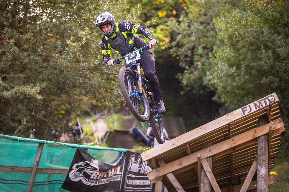Mountain biker in black and green gear mid-jump off ramp in forest trail. Wooden platform labeled "FJ MTB." Energetic and focused.