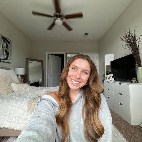 Smiling Grace takes a selfie in a cozy bedroom. Gray hoodie, wood fan, white bedding, mirrored closet, and dresser in the background. Relaxed vibe.
