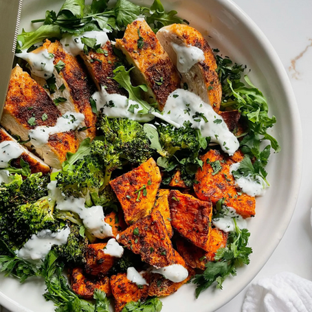Grilled chicken, roasted sweet potatoes, and broccoli on greens, drizzled with white sauce on a white plate. Fresh, vibrant, and appetizing.