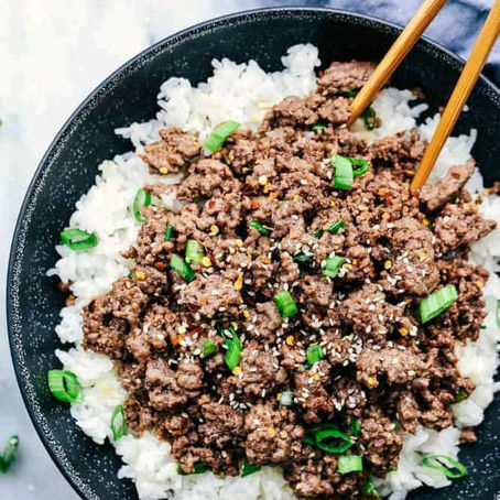A black bowl with rice topped with seasoned ground beef and green onions. Chopsticks are placed on top. Marble surface in the background.