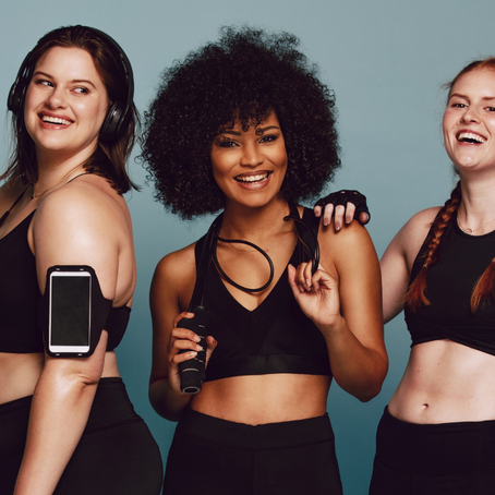 Three women in black workout gear smile joyfully against a blue background. One holds a jump rope, another wears headphones and a phone armband.