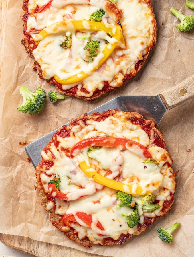 Two mini vegetable pizzas with melted cheese, bell peppers, and broccoli on parchment, one resting on a spatula.
