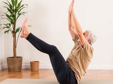 Elderly man doing yoga boat pose on a mat in a bright room with wooden floor. Potted plant in background. Calm and focused expression.