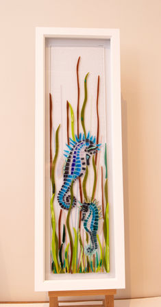 Transform your living space with this beautiful one-off piece of fused glass art by artist Greig Black. The artwork depicts two enchanting blue sea horses gracefully holding onto sea grass, meticulously crafted using a stunning combination of transparent, opaque, and dichroic glass. This exclusive creation embodies the unique handmade quality and personalized touch that blacknoir glass is celebrated for. Perfect for adding a touch of marine elegance to any wall, this piece is not just art—it's a statement of individuality and craftsmanship. Make it yours and bring the ocean's charm into your home. The piece is 52.5cm x 13cm and is framed in a white box frame 60cm x 20cm.
Cost: £250.00