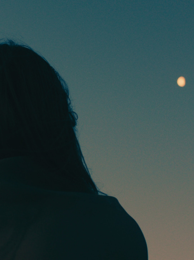 The back of a woman at dusk looking up at a full moon against a teal sunset.