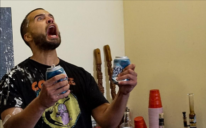 Film still of the character Justen holding drinks and reacting energetically inside an interior setting.