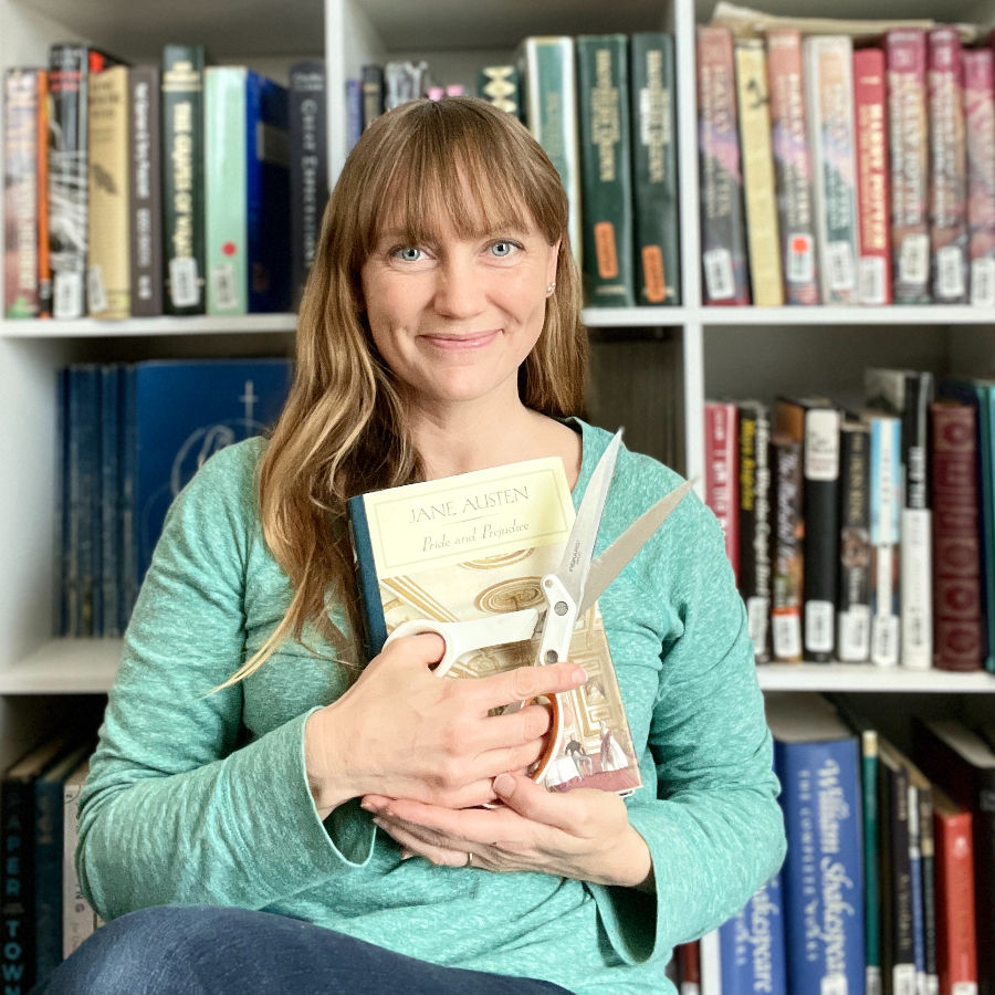 Katie Lloyd, owner and designer at Anthology On Main, sitting in front of a library of books, holding scissors and a hardcover copy of Pride and Prejudice by Jane Austen, ready to be turned into beautiful and long lasting book page flowers 