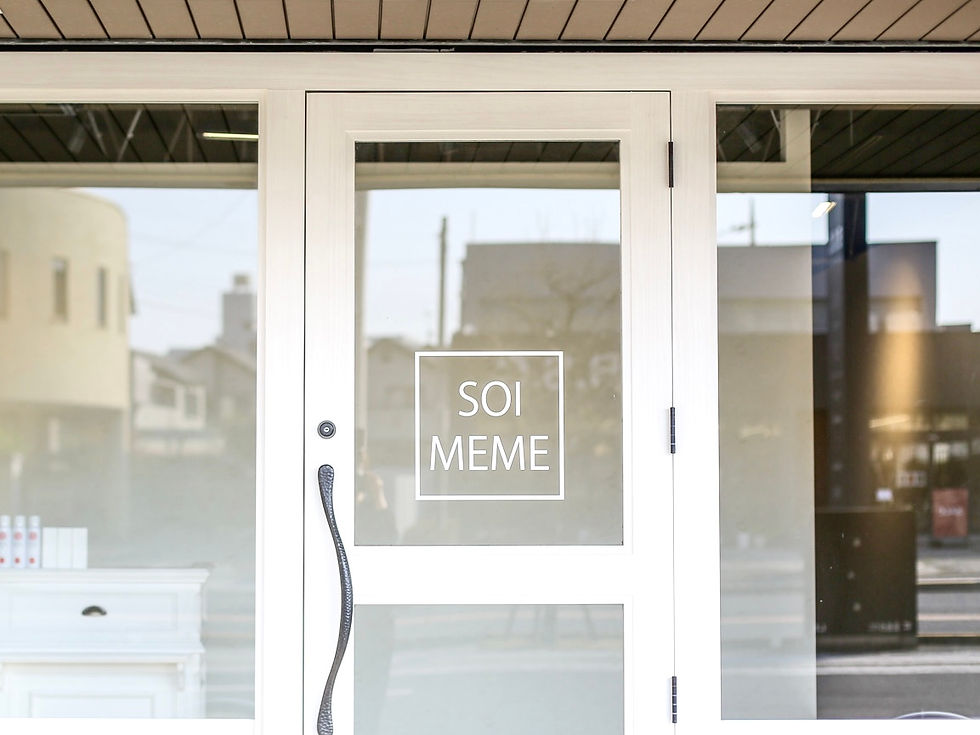 白いドアに「SOI MEME」と書かれた看板。ガラス越しに街並みが反映され、店内に白い家具が見える。明るくモダンな雰囲気。