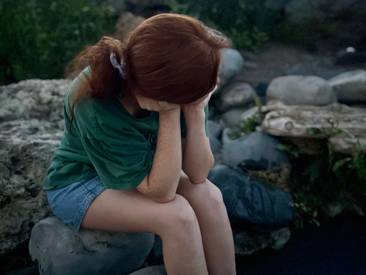 a woman rests alone on a rock in the middle of a brook. She is pensive, thoughtful and sullen. Image feels dark and overwhelmed. 