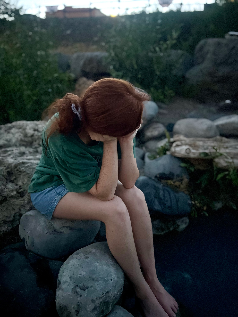 a woman rests alone on a rock in the middle of a brook. She is pensive, thoughtful and sullen. Image feels dark and overwhelmed. 