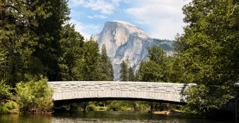 Yosemite half dome is slightly blurred in the background with a rock bridge in the foreground, framed by ever green trees and a blue sky with scattered clouds. There is a meandering river that the bridge spans and the waters are tranquil and reflective.