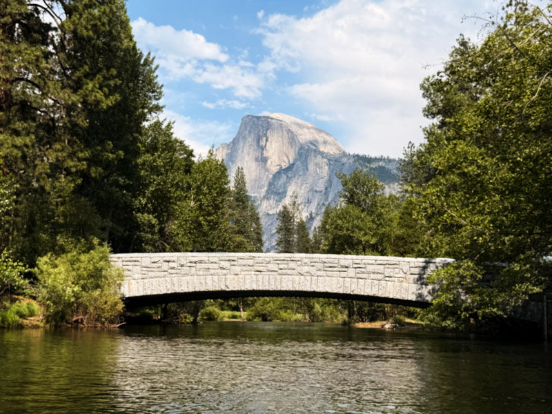 Yosemite half dome is slightly blurred in the background with a rock bridge in the foreground, framed by ever green trees and a blue sky with scattered clouds. There is a meandering river that the bridge spans and the waters are tranquil and reflective.