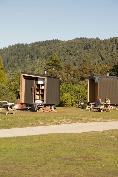 Two modern cabins on trailers, people enjoying picnic tables outdoors near forest