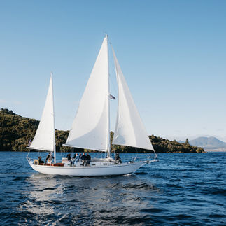White-sailed yacht cruising past lush green land on a calm day in Lake Taupō.