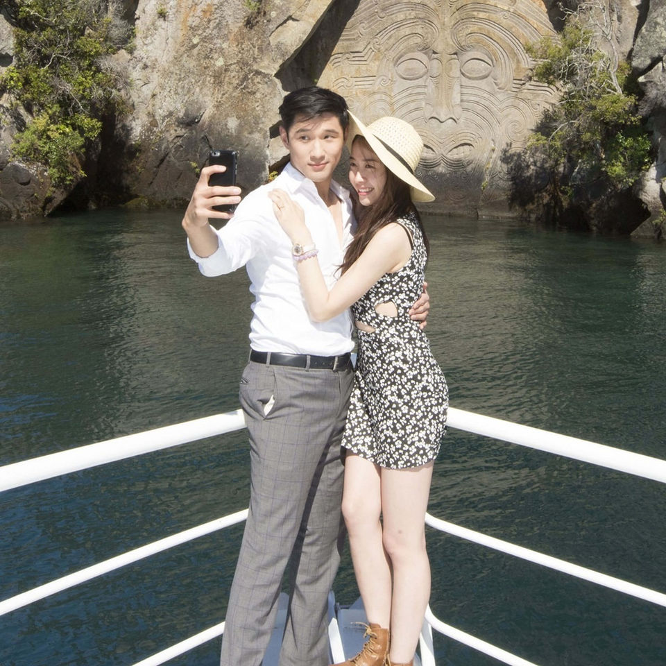 Couple taking a photo aboard a Sail Barbary sailing cruise with the Māori Rock Carvings in the background.