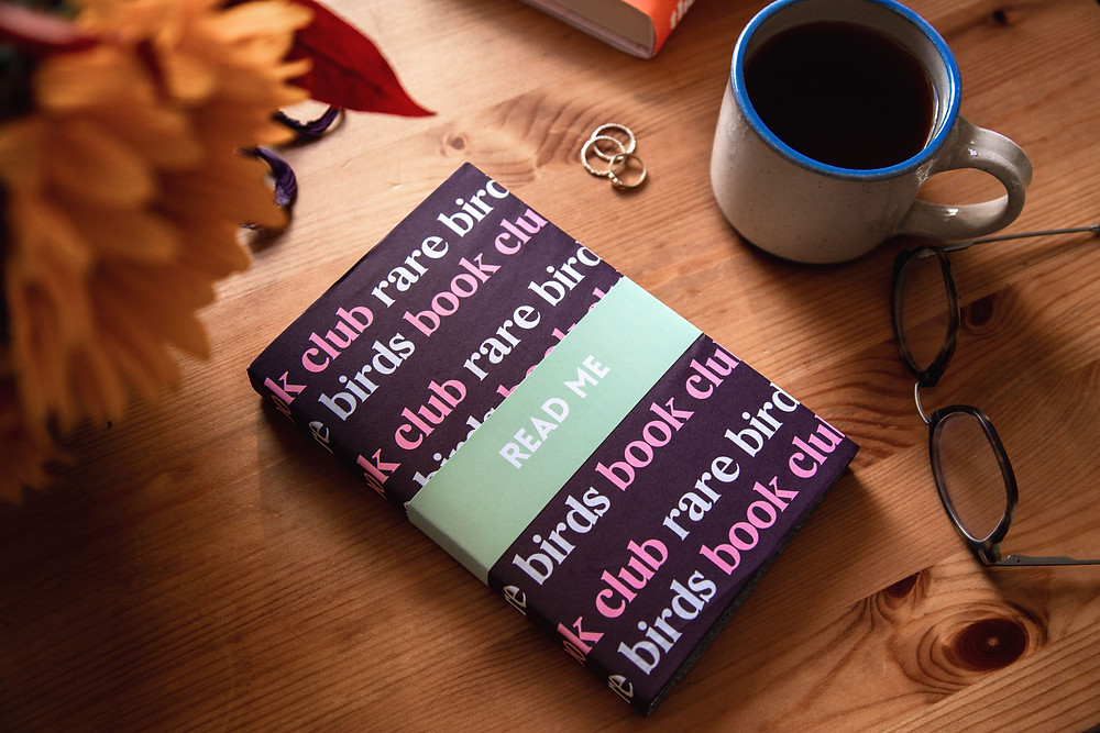 A photo of a Rare Birds Book Club book, laid on a desk next to a pair of glasses and a mug of black coffee.