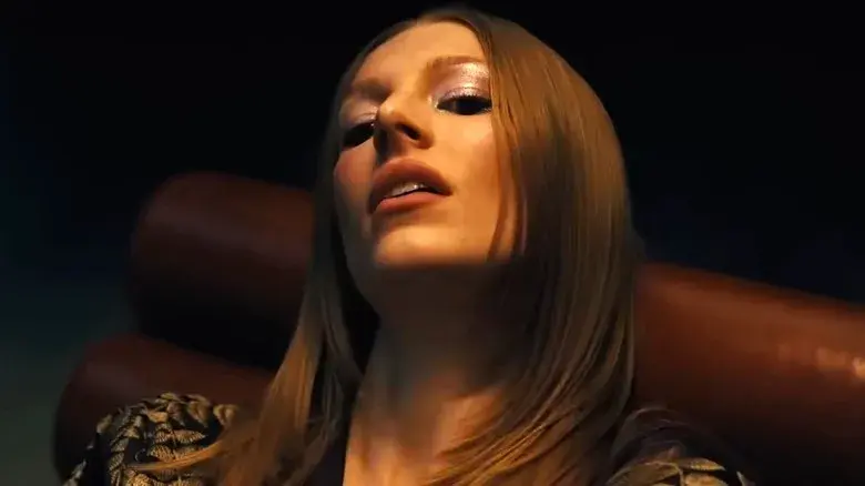 Close-up of a woman with long straight hair reclining in a brown leather chair, lit warmly against a dark background, moody expression