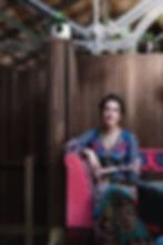 Woman in maximalist playsuit sat in a pink velvet chair with patterned throw. Industrial ceiling beams and wood panelled wall background