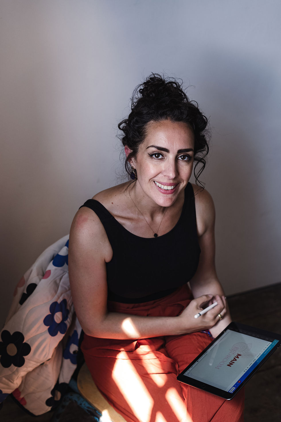 Woman smiling, sitting on a stool with a tablet showing design plans. Wears black top, red trousers. Floral coat on chair, light shadows.