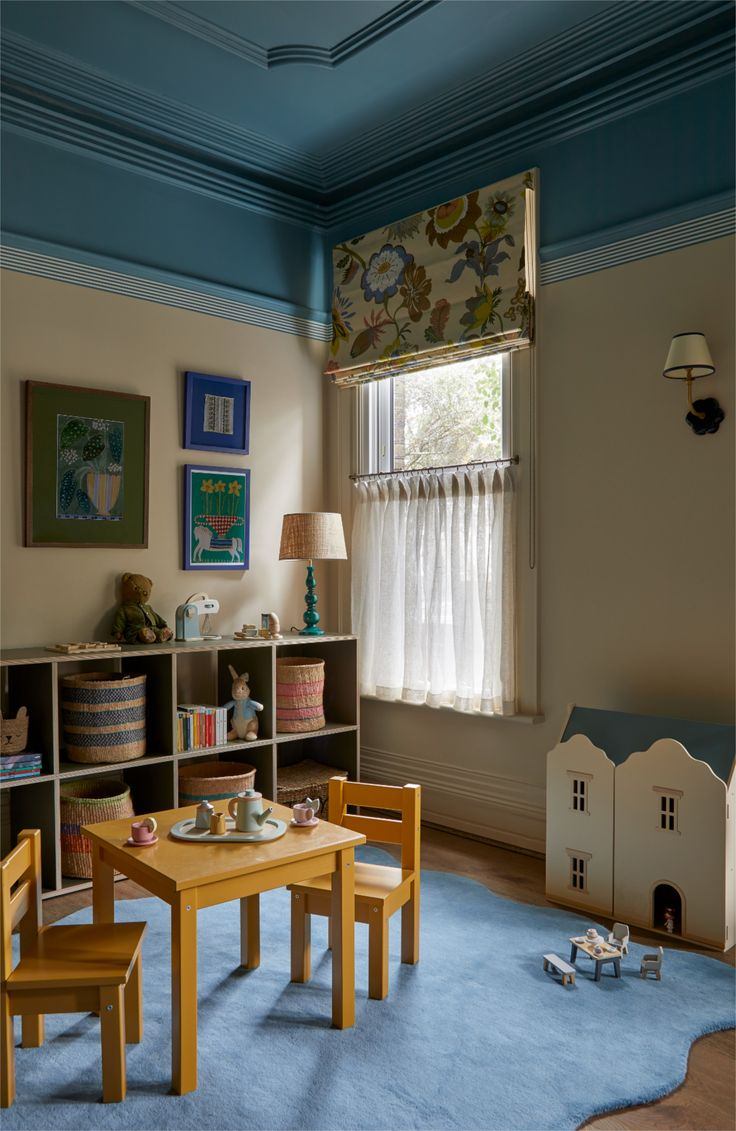 Whimsical children’s playroom with yellow chairs, tea table, toy house, stuffed animals, blue rug, floral blind, and cubby shelves