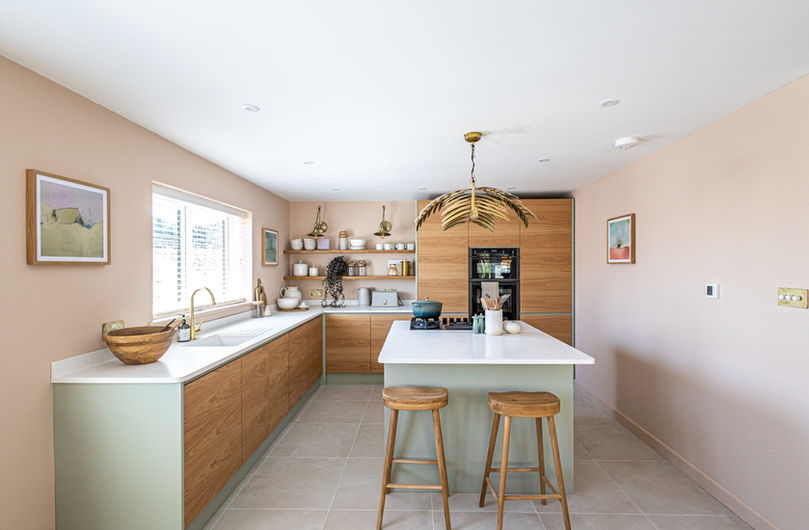 Bespoke wooden kitchen with minerva worktops, hob with extra fan built in for seamless design. abstract art and brass accessories in the kitchen with a stone floor. Green and pinks blended together for a bold coastal interior design.