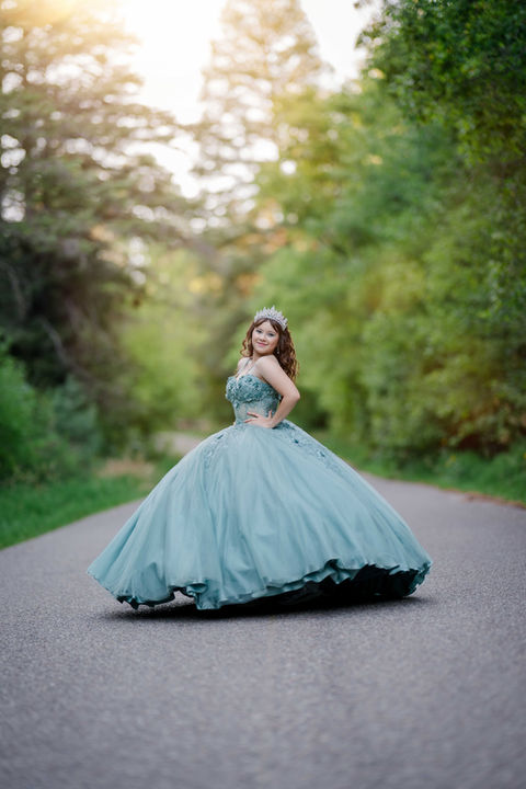 Quinceañera in a teal ball gown posing on a tree-lined outdoor pathway.
