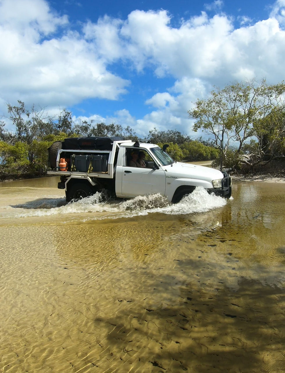 Landscape photo of a white 4WD ute driving through shallow water under a bright blue sky with clouds and trees in the background. The vehicle is centered, showing off-road capability and adventure. Aspect ratio: portrait.