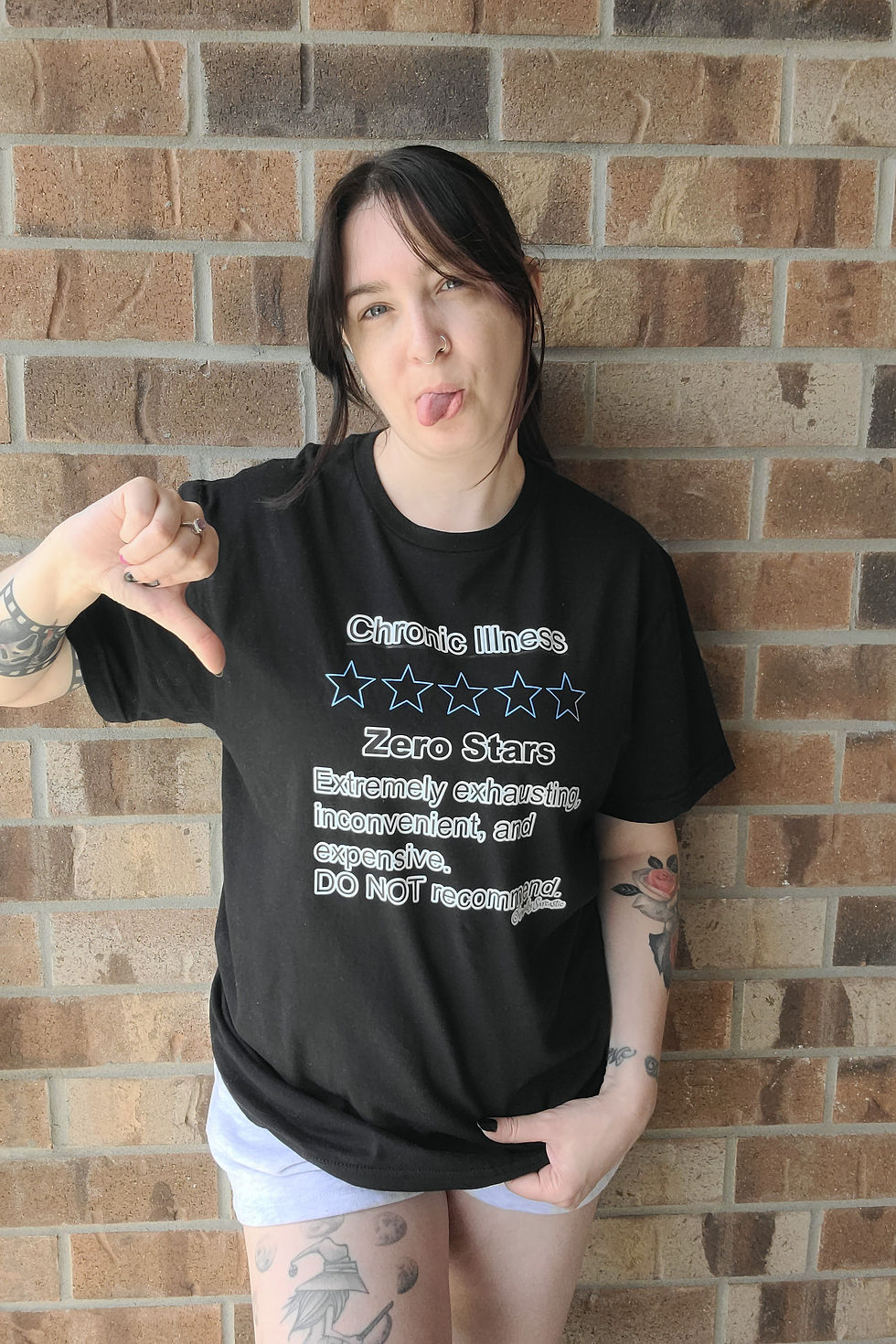 Girl wearing a black t-shirt with "Chronic Illness Zero Stars" funny awareness design on a brick wall with her thumb down
