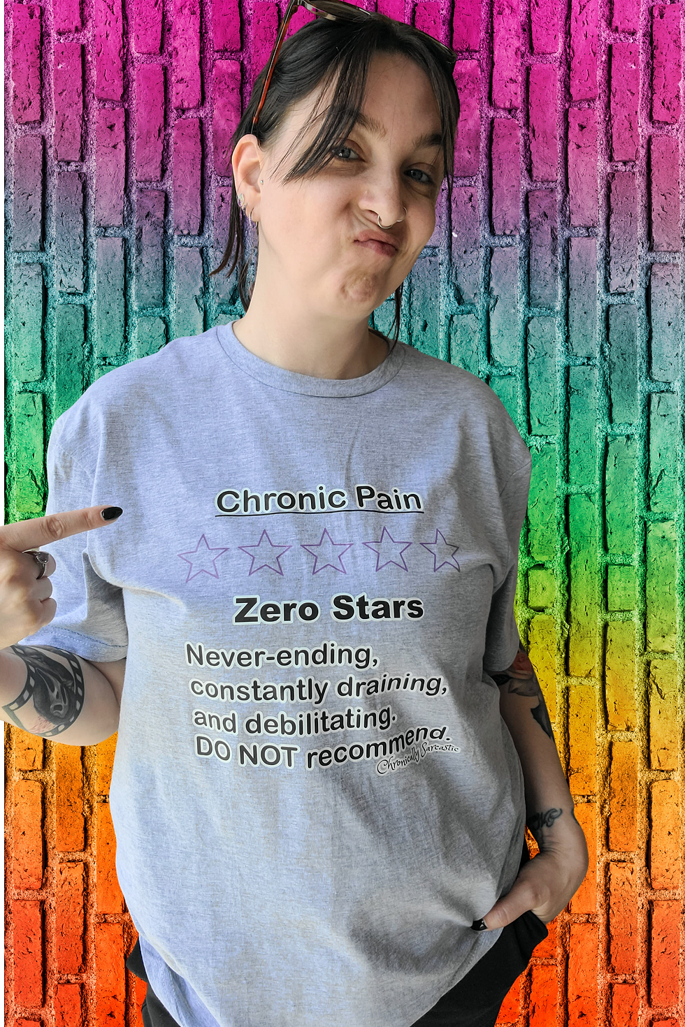 Girl standing on a pride wall wearing a grey tee with " Chronic Pain 0 Stars" funny review design pointing at the design