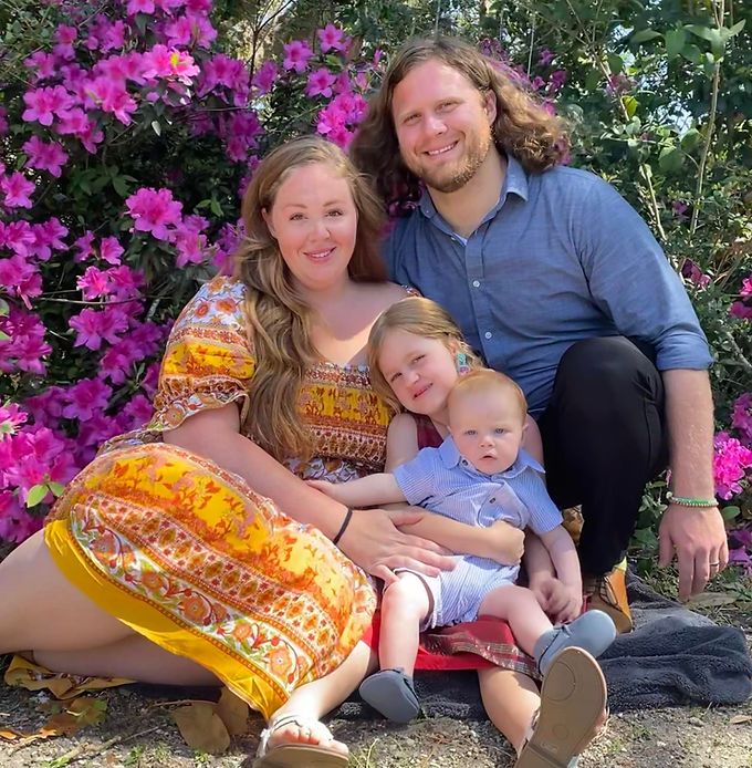 Young man with long hair, wife and two small children sit on the ground, in front of an Azalea bush.