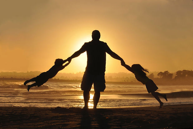 A man standing on a beach is swinging a child from each arm at sunset.