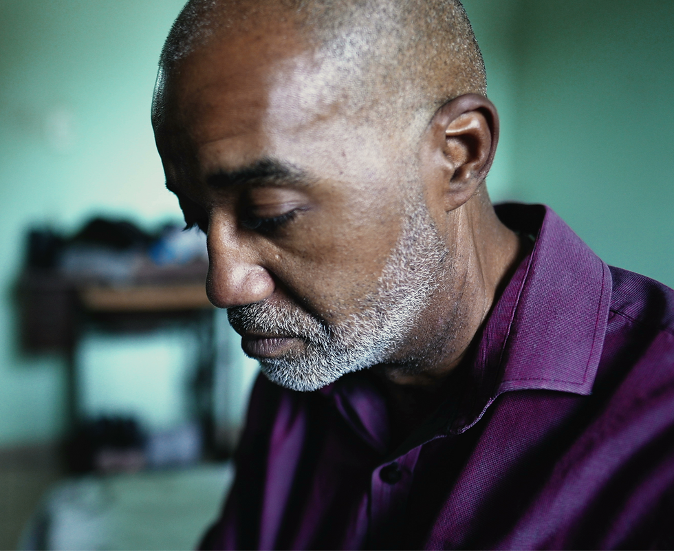 An older black man with a short gray beard and a purple shirt. He looks sad, sitting with his back hunched and his face downturned.
