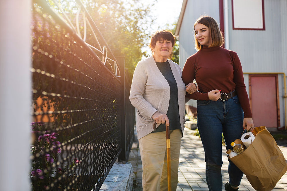 A younger woman in a red top stands arm-in-arm with an older woman in a gray sweater while carrying a bag of groceries. They are both smiling and standing outside in the sunshine beside a reflective wall.
