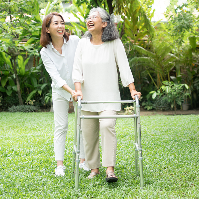 A young, smiling Asian woman with brown hair stands partly behind and older Asian woman with gray hair. The older woman is using a walker do you move across a green lawn while the Younger woman assists her. There is lush tropical landscaping in the background.