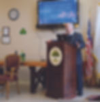 A balding white man with gray hair and a gray mustache is standing behind a dark wooden podium in a room with beige walls and a beige linoleum floor. He is speaking while a blue slide with white text is displayed on a monitor mounted to the wall behind him. There is an American flag in a stand in the corner behind him, a bulky wooden table to his left, and an emblem with a tree in the center on the front of the podium. The man is wearing a dark sweater over a white collared shirt with a blue tie, dark pants, black shoes, and a dark charcoal jacket.